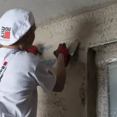Pracownik w białej czapce i rękawiczkach nakłada tynk silikonowy. Kluczowe jest, po jakim czasie zacierać tynk silikonowy, by uzyskać idealną gładkość.