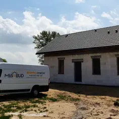 Biała furgonetka budowlana M&S BUD przed domem w budowie z szarym dachem i oknami.