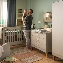 Ojciec trzyma dziecko w pokoju niemowlaka. Wnętrze pokazuje, jak urządzić pokój niemowlaka: łóżeczko, przewijak, szafę i fotel.