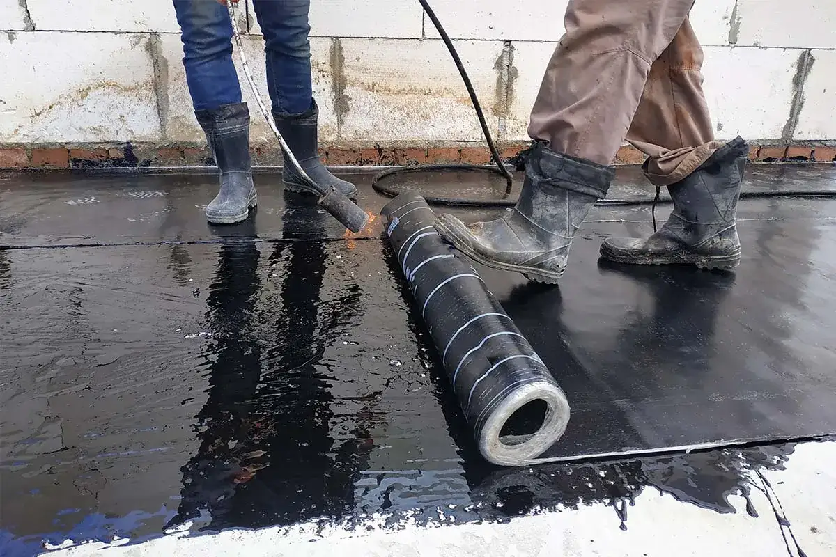 Dwóch robotników układa lepik na dachu, używając palnika do rozgrzewania materiału.