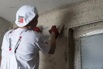 Pracownik w białej czapce i rękawiczkach nakłada tynk silikonowy. Kluczowe jest, po jakim czasie zacierać tynk silikonowy, by uzyskać idealną gładkość.