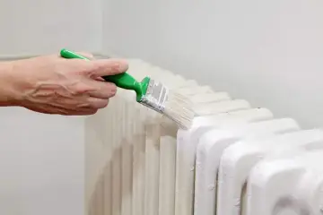 Ręka z zielonym pędzlem odnawia biały grzejnik. Malowanie grzejnika to sposób na odświeżenie wnętrza.